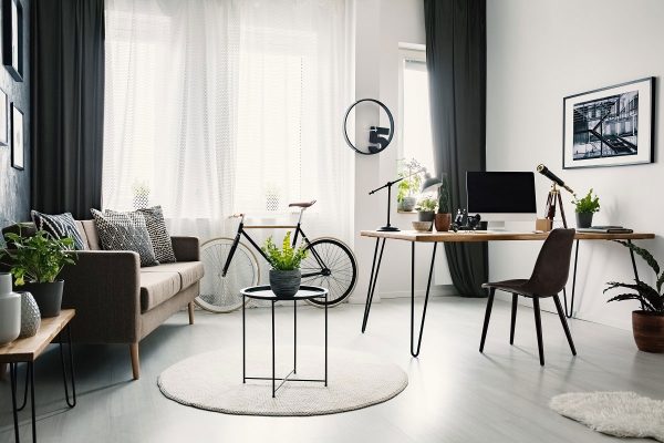 Bright living room interior with sofa with cushions, bike under the window with curtains and hairpin desk with lamp and computer for remote work